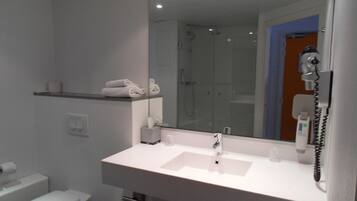 Un baño moderno con lavamanos blanco, espejo y retrete. Hay un perchero sobre el lavamanos y una ducha visible en el espejo.