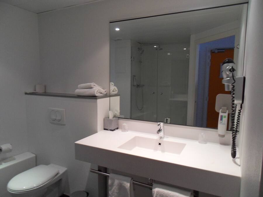 Un baño moderno con lavamanos blanco, espejo y retrete. Hay un perchero sobre el lavamanos y una ducha visible en el espejo.