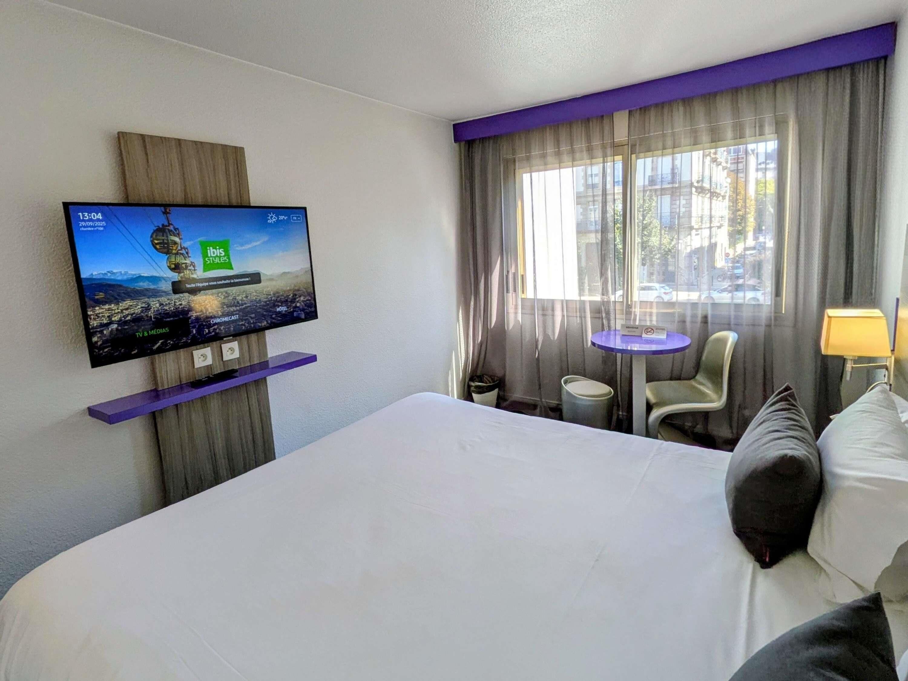 Quarto de hotel com cama, televisão presa a um painel de madeira, mesinha, cadeira e janela com cortinas.