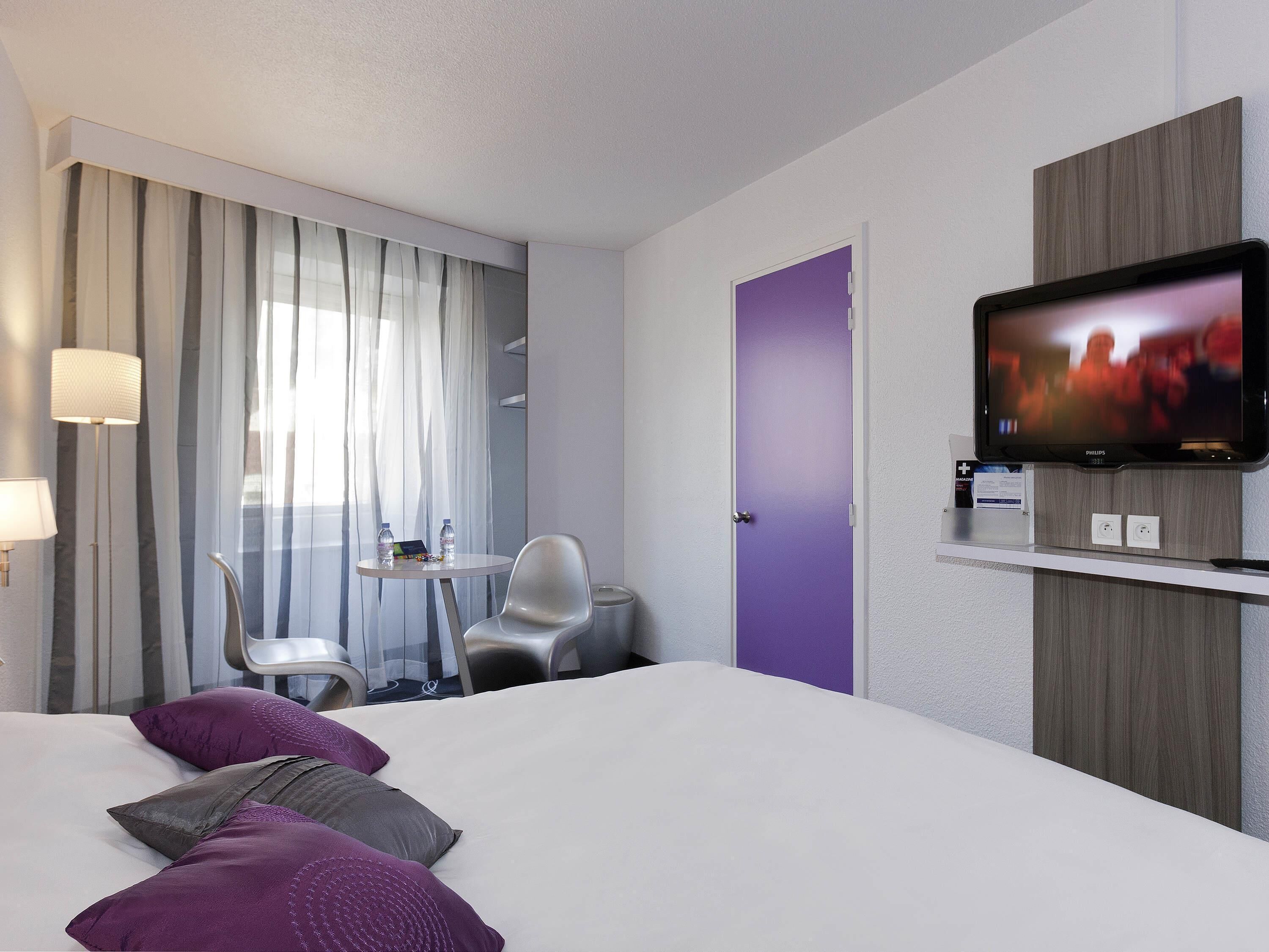 Ein Hotelzimmer mit Bett, Fernseher, Schreibtisch mit Stuhl, Fenster mit Vorhängen und einer Tür.