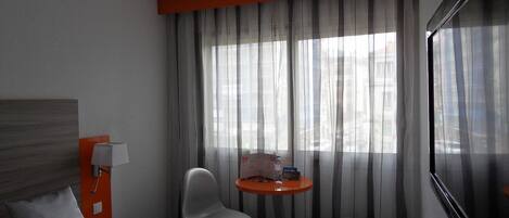 A hotel room with a bed, a chair, a small table with a bottle and a glass, and a window with curtains.