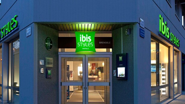 L'entrée d'un hôtel ibis Styles, avec des portes en verre, une enseigne au-dessus de l'entrée et un écran numérique sur le mur.