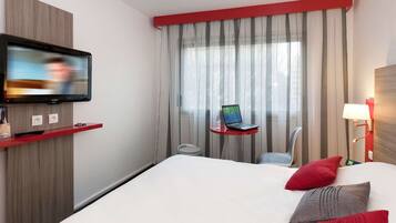 Habitación de hotel con una cama, un televisor montado en la pared, un ordenador portátil sobre una mesita y una lámpara.