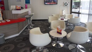 Un espace d’accueil d’hôtel avec un comptoir rouge, des chaises modernes, un téléviseur et une moquette à motifs.