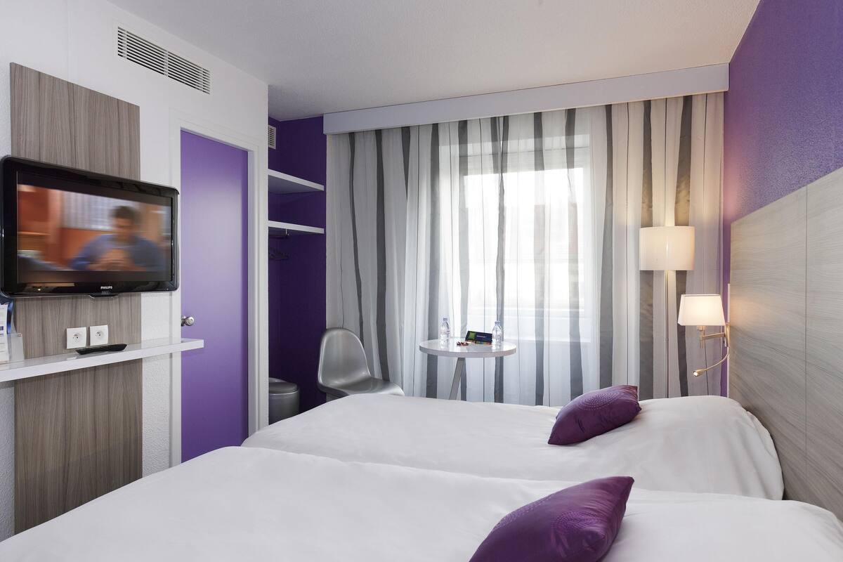 Habitación de hotel con televisión, dos camas, una silla, una mesita y una ventana con cortinas.