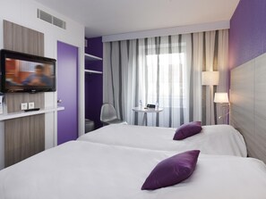 Een hotelkamer met een televisie, twee bedden, een stoel, een klein tafeltje en een raam met gordijnen.