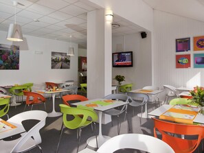 Een moderne eetruimte met witte tafels, kleurrijke stoelen en een televisie aan de muur.
