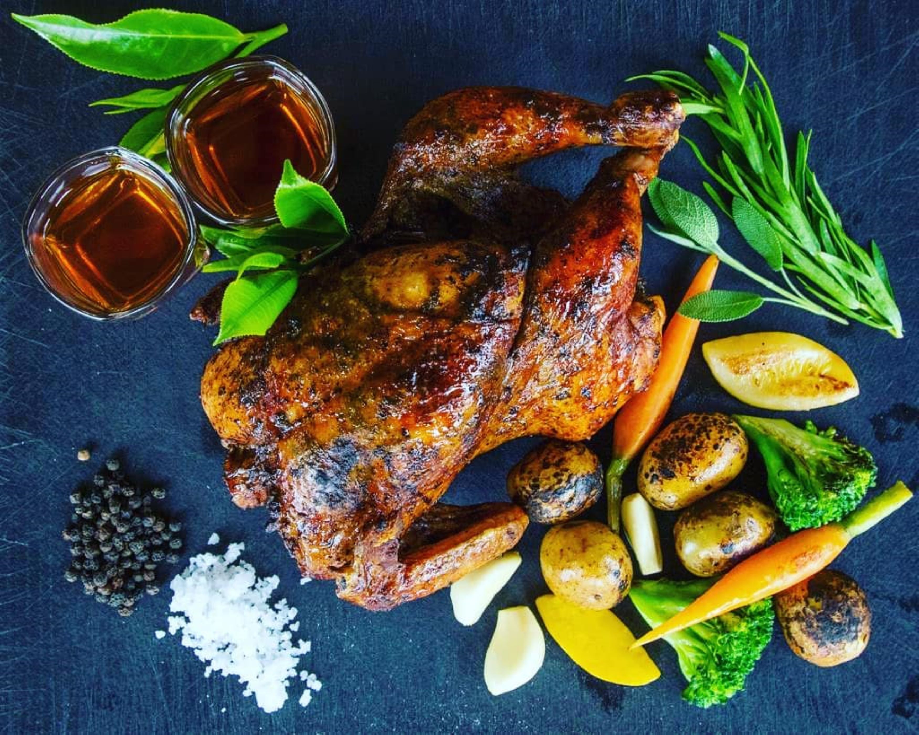 Pollo asado con vegetales y hierbas sobre una superficie oscura.