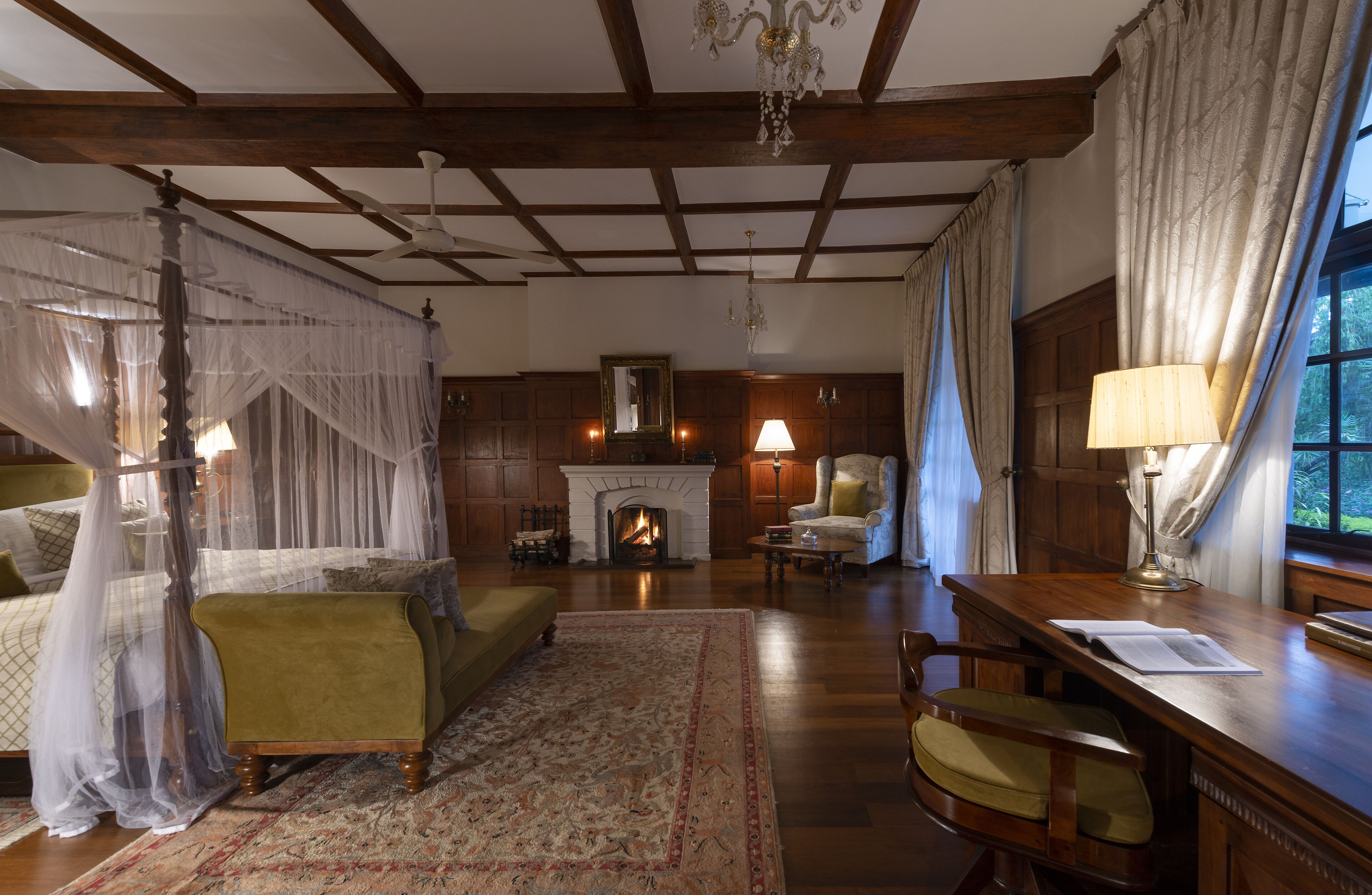 A traditional room with a four-poster bed, a fireplace, a desk with a lamp, and a view of the outdoors.
