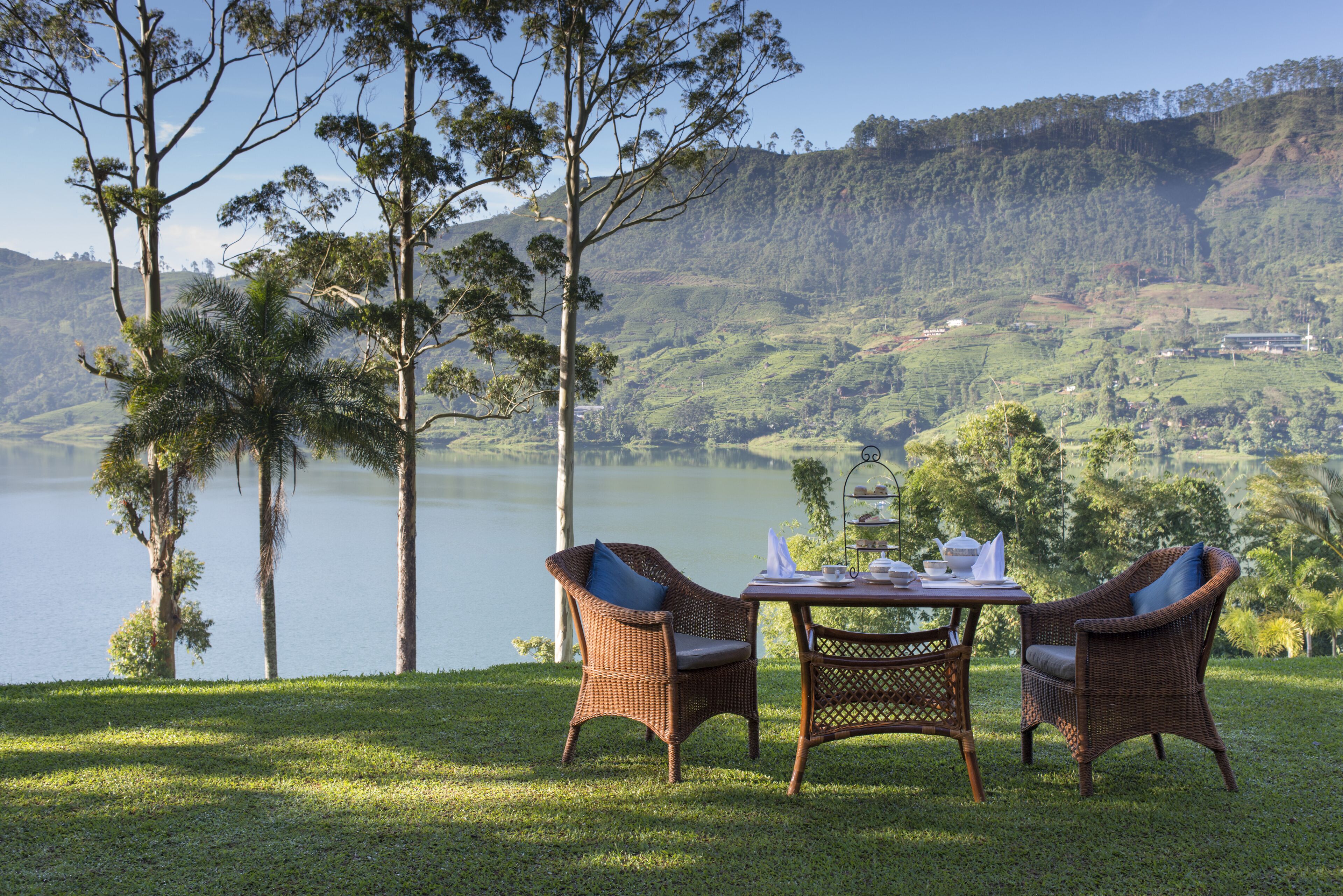 Un coin repas avec des meubles en osier et une table dressée pour deux personnes, offrant une vue sur un lac et des collines.