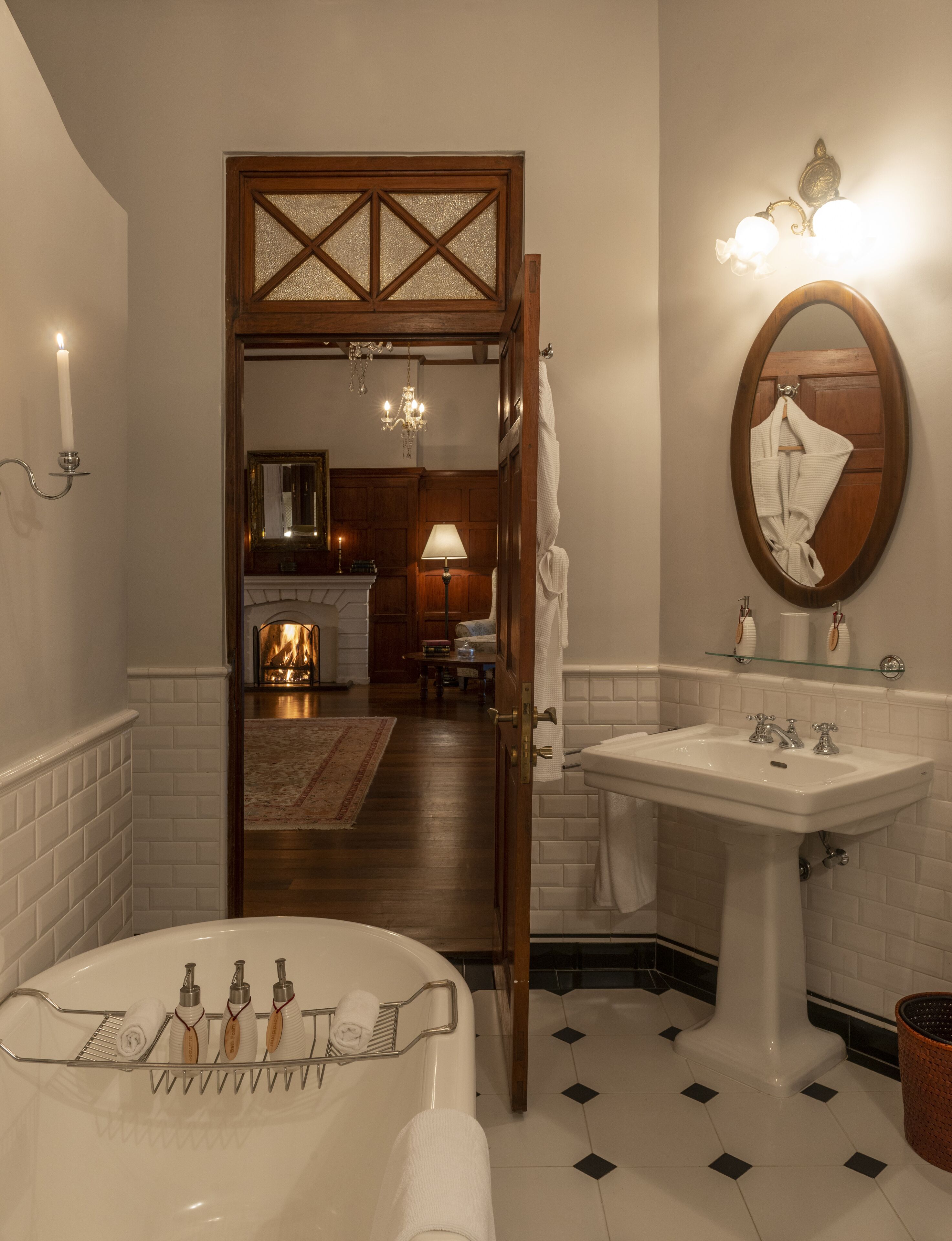 Ein Badezimmer mit einer Badewanne, einem Waschbecken und einem Spiegel. Eine Tür führt zu einem Wohnbereich mit Kamin.