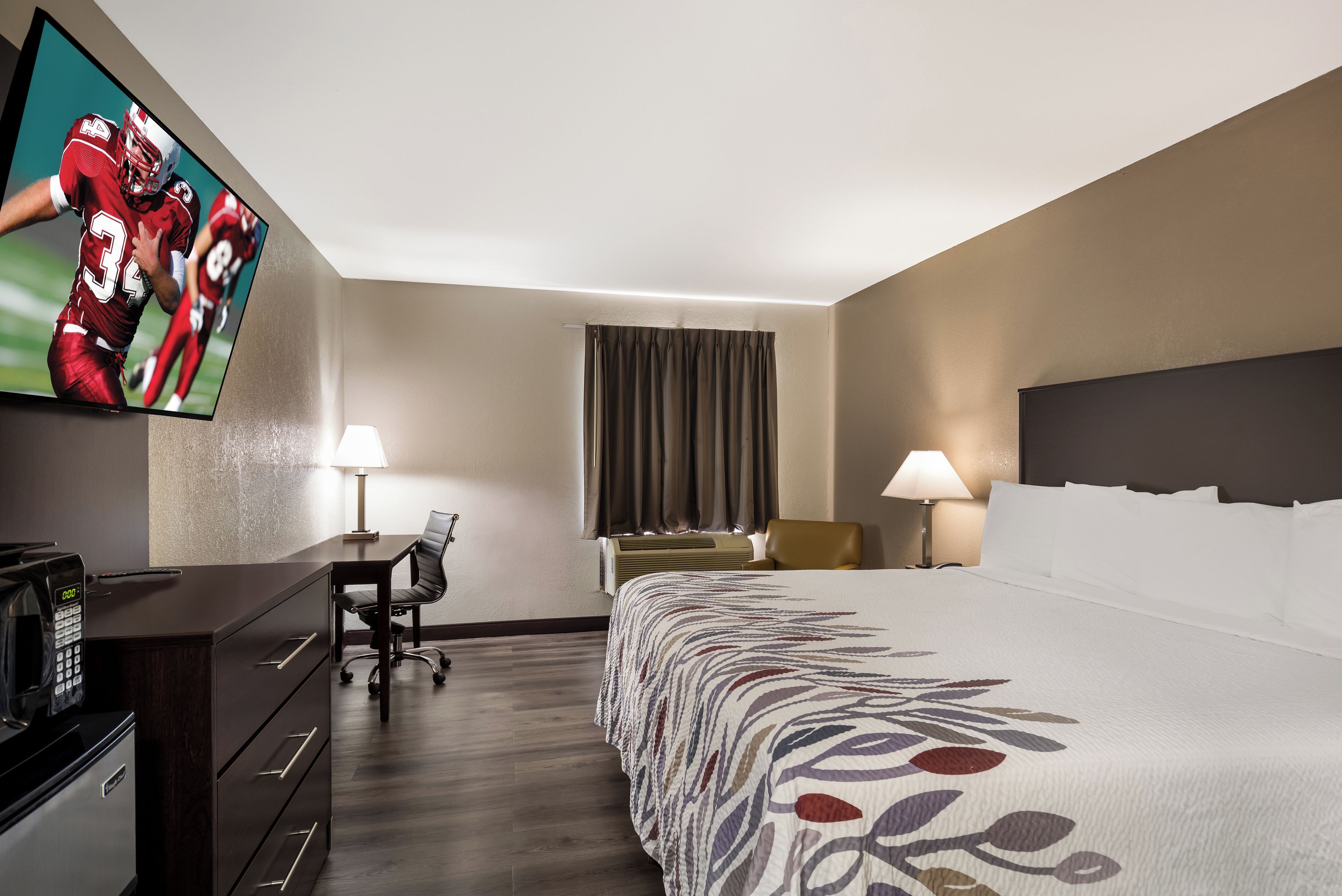 Una habitación de hotel con cama, escritorio, silla, televisor y ventana con cortinas.