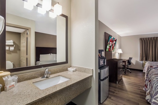 Habitación de hotel con lavamanos de encimera de granito, refrigerador, televisor de pantalla plana, cama con cubrecama estampado y escritorio con silla.