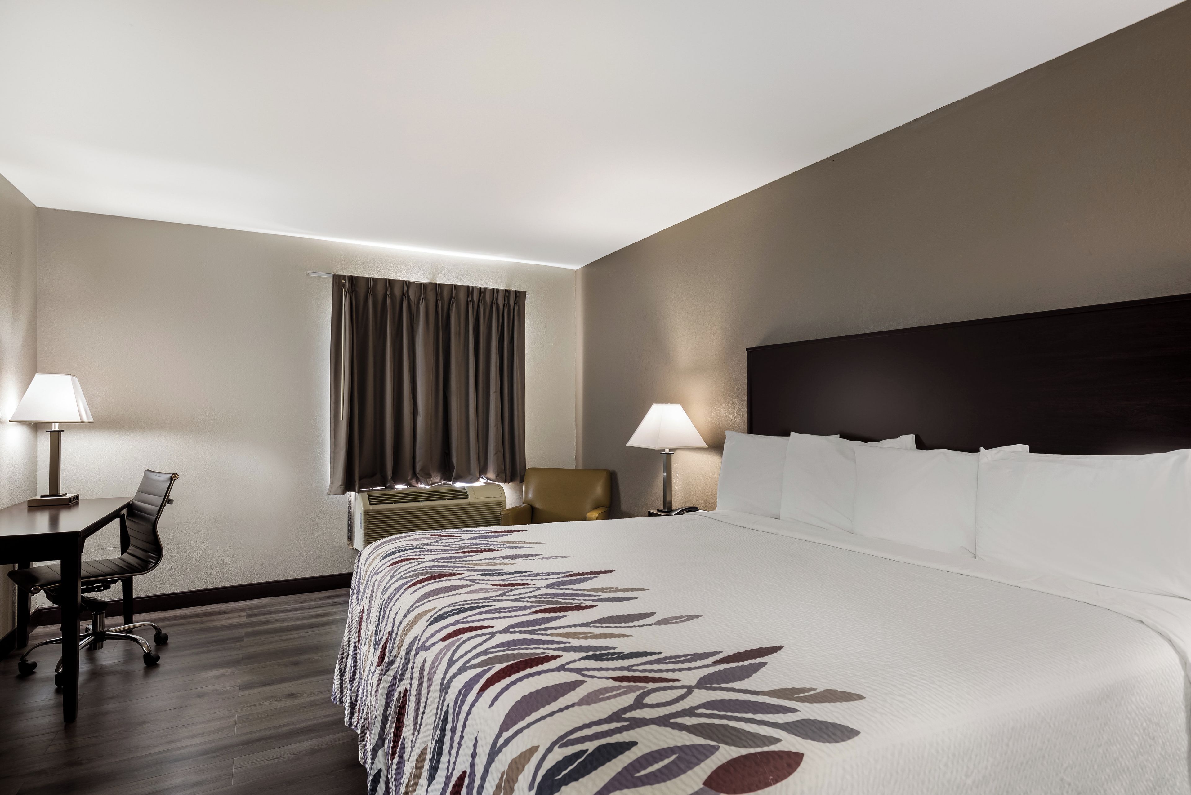 A hotel room with a large bed, two bedside lamps, a desk with a chair, and a window with curtains.