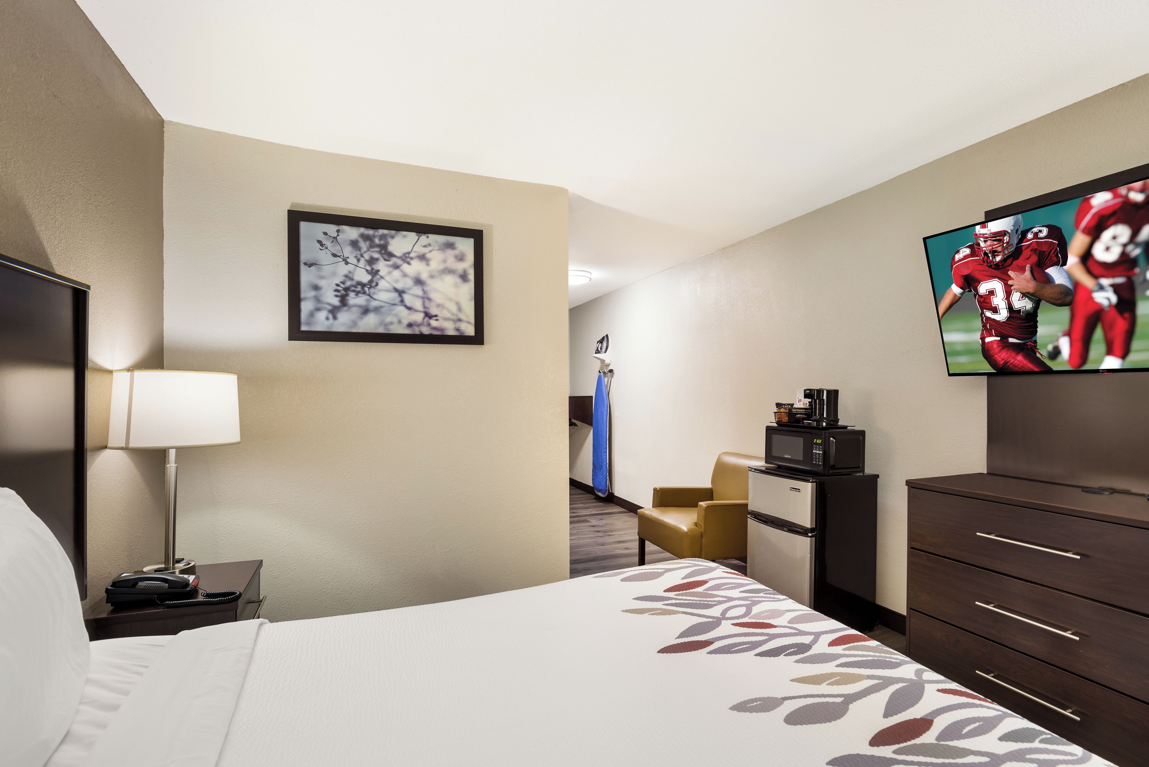 A hotel room with a bed, a desk with a lamp, a television, a minibar, and a seating area with a chair.