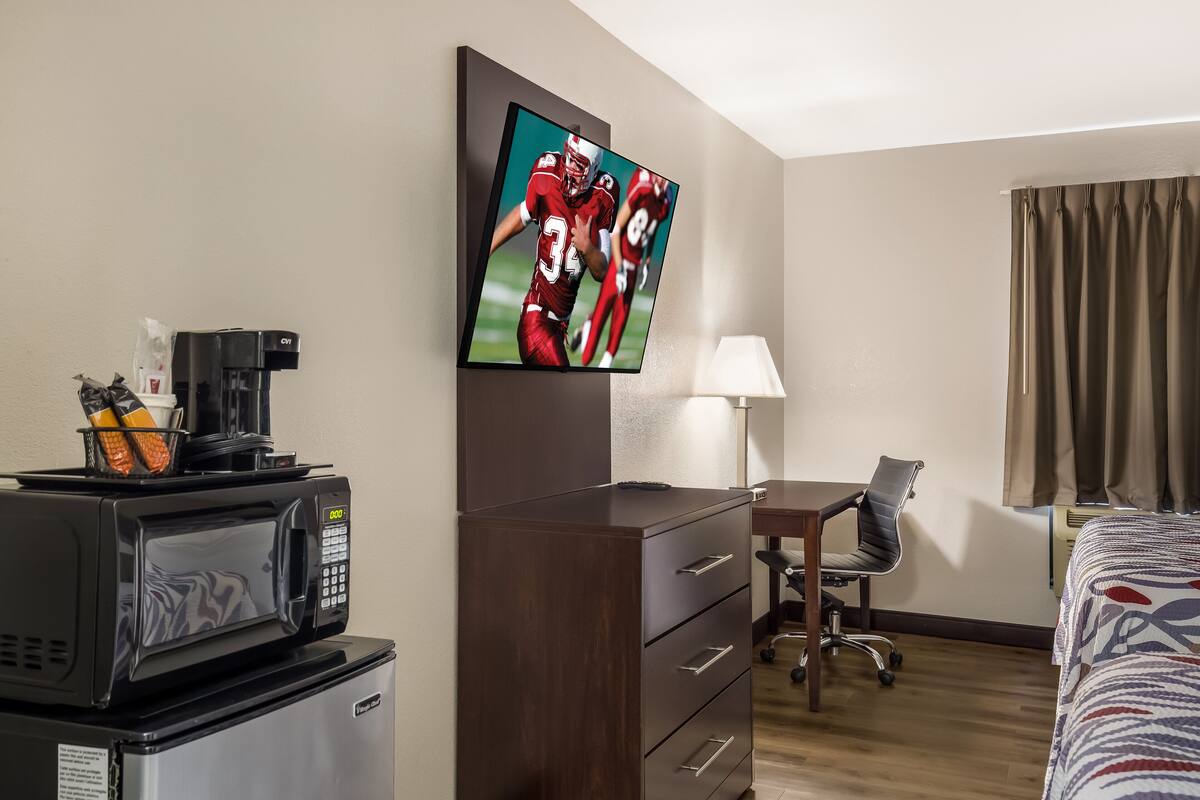 A hotel room with a flat-screen TV, a desk, a chair, a bed, a coffee maker, a microwave, and a refrigerator.
