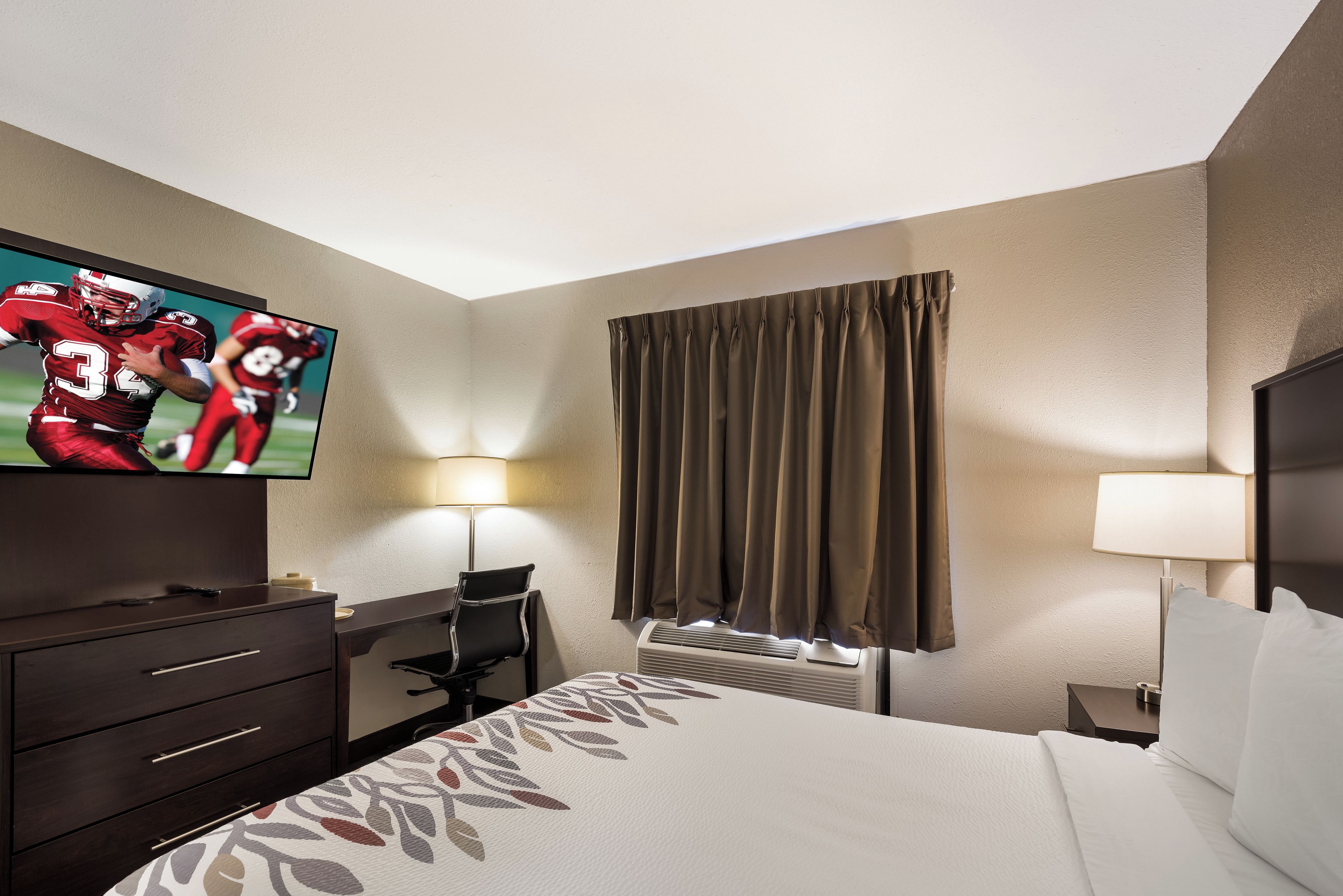 A hotel room with a bed, a desk, a television, a lamp, and a window with curtains.