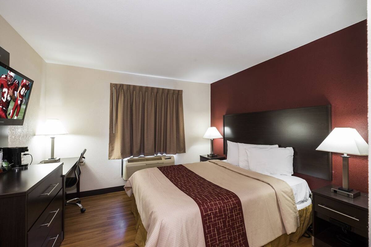 A hotel room with a bed, bedside tables, a desk, a chair, a TV, and a window with curtains.