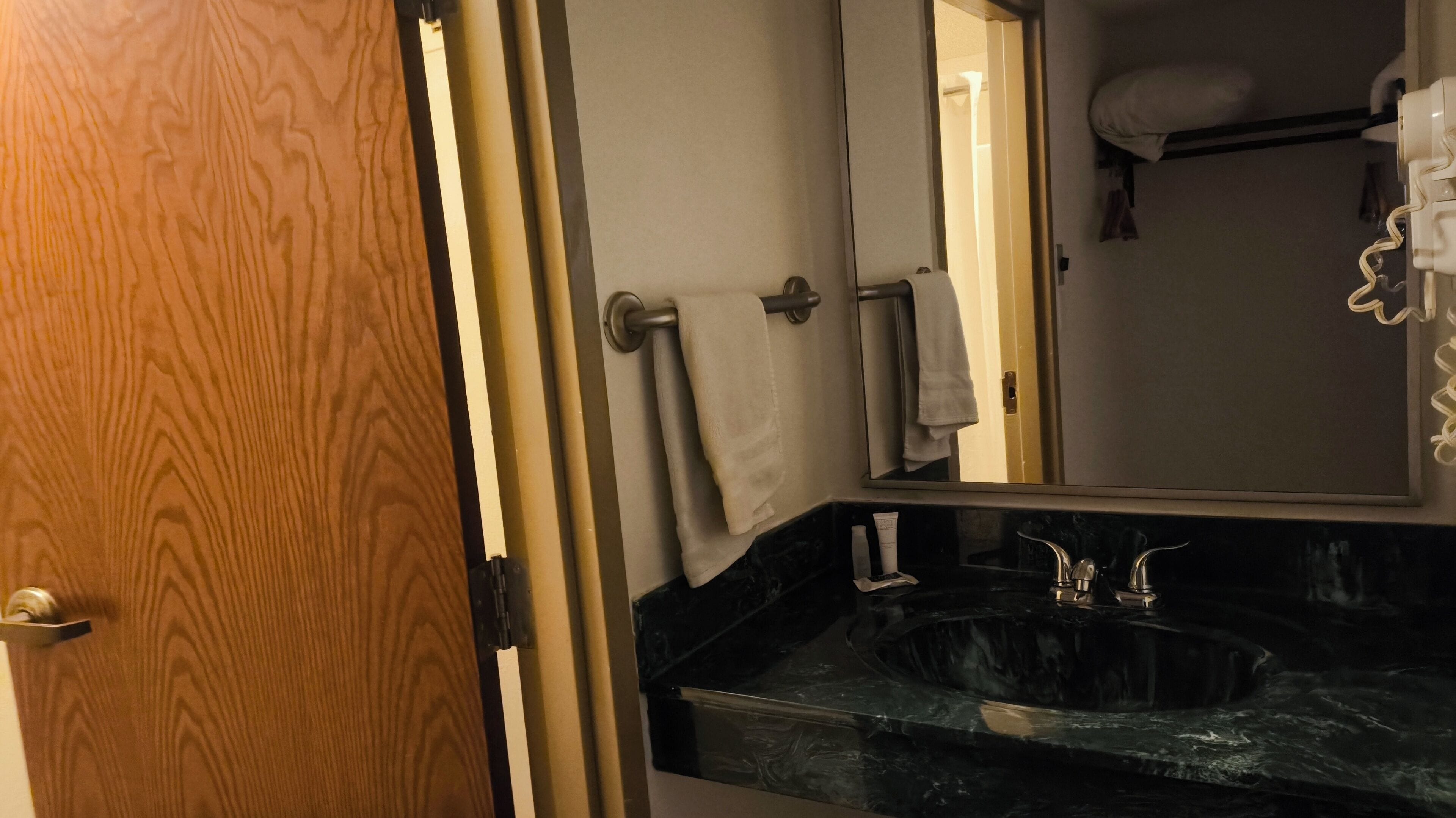 A bathroom with a wooden door, a towel rack, a sink with a marble countertop, and a mirror.