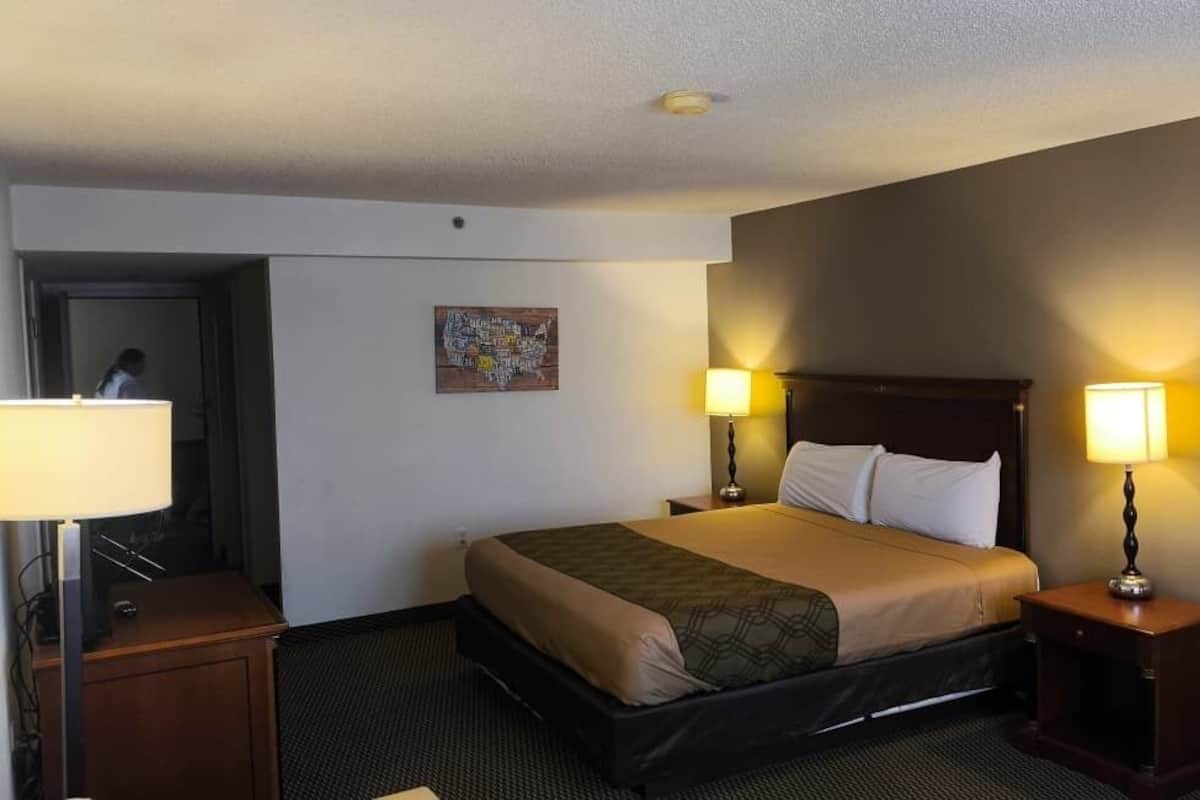 A hotel room with a bed, two bedside lamps, a wooden headboard, a door, and a small table with a lamp.
