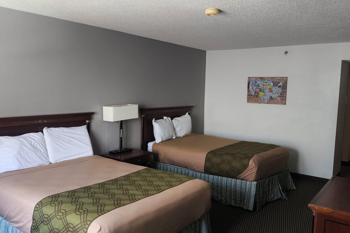 A hotel room with two beds, a nightstand with a lamp, a painting on the wall, and a door to the right.