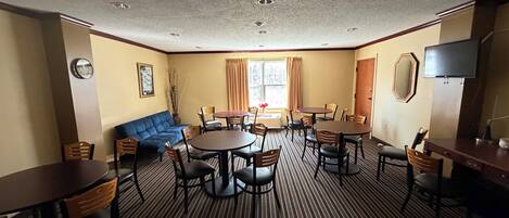 A hotel room with a blue sofa, multiple dining tables, chairs, a TV, and a large window with curtains.