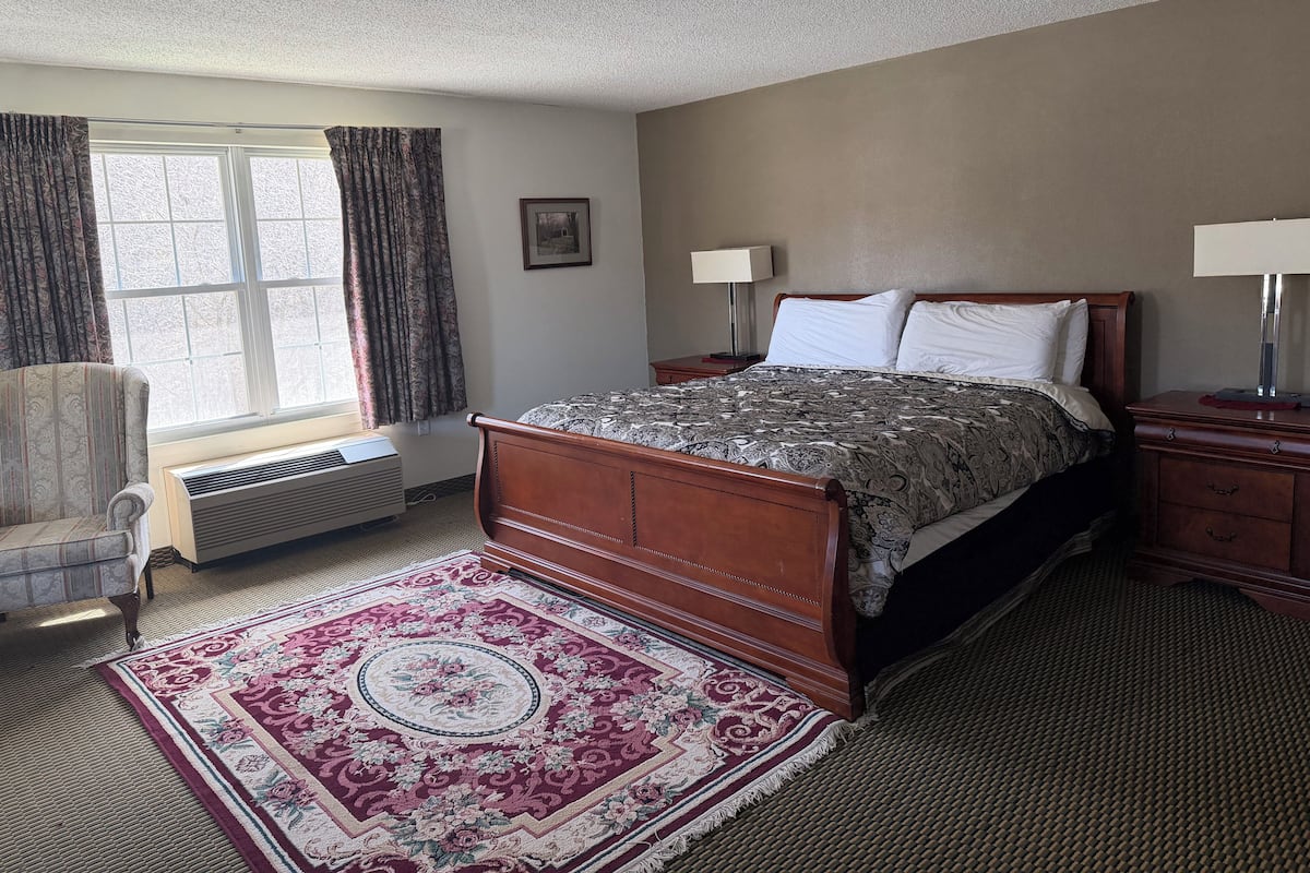 A bedroom with a bed, two bedside tables, a chair, a window with curtains, a radiator, and a rug.