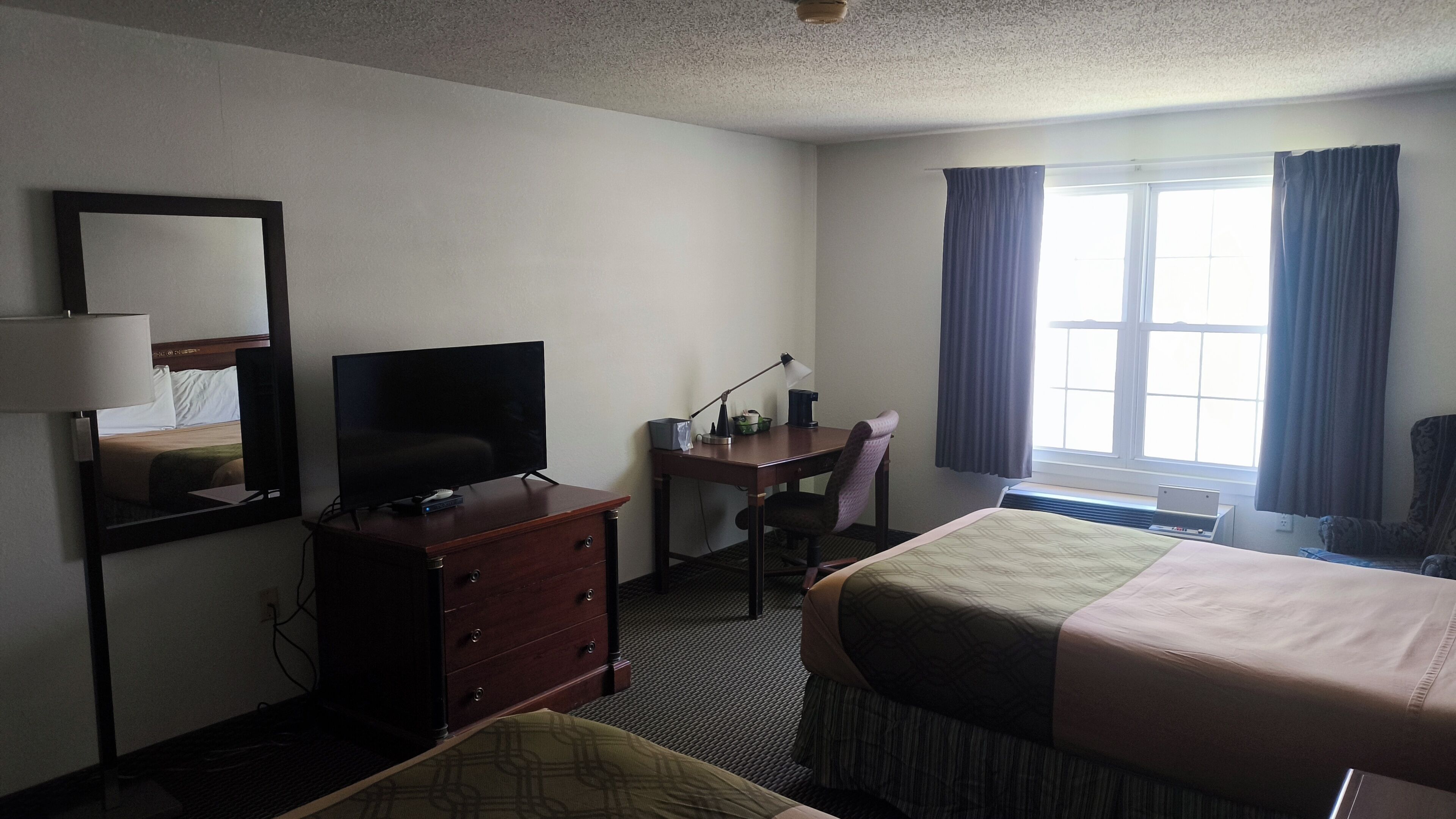 Habitación de hotel con cama, escritorio, silla, televisor, cómoda, espejo y ventana con cortinas.