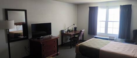Habitación de hotel con cama, escritorio, silla, televisor, cómoda, espejo y ventana con cortinas.