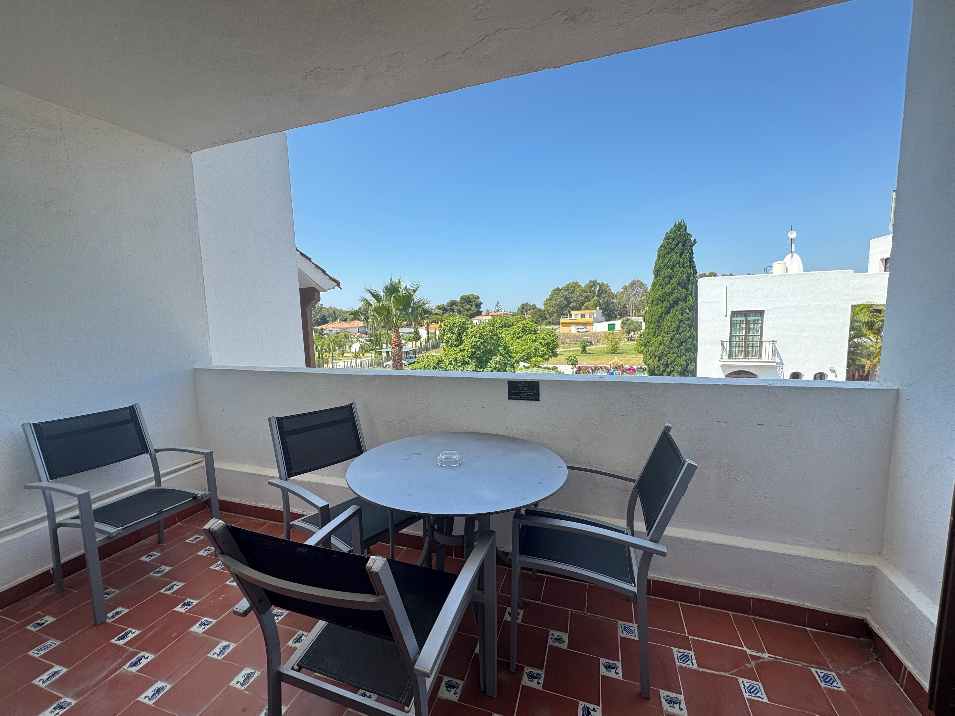 Un balcon avec une table ronde et quatre chaises, un sol carrelé et une vue sur des immeubles et des arbres.