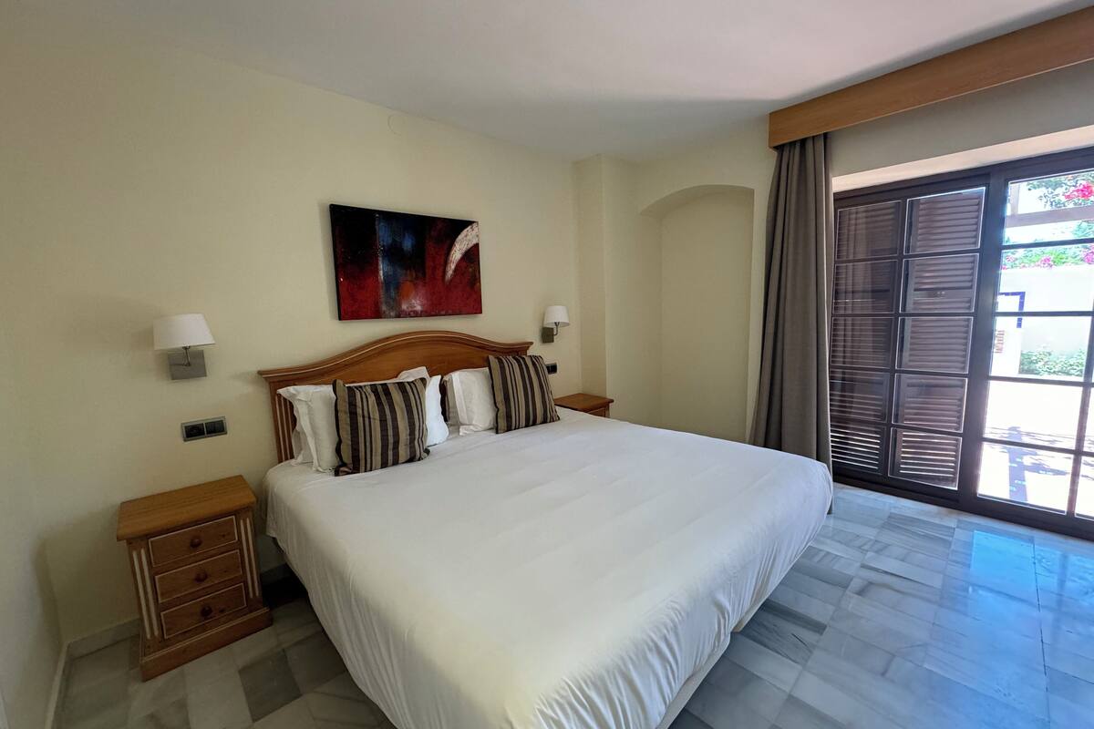 Ett hotellrum med en stor säng, två sängbord, ett litet bord, en tavla på väggen och utsikt genom fönstret.