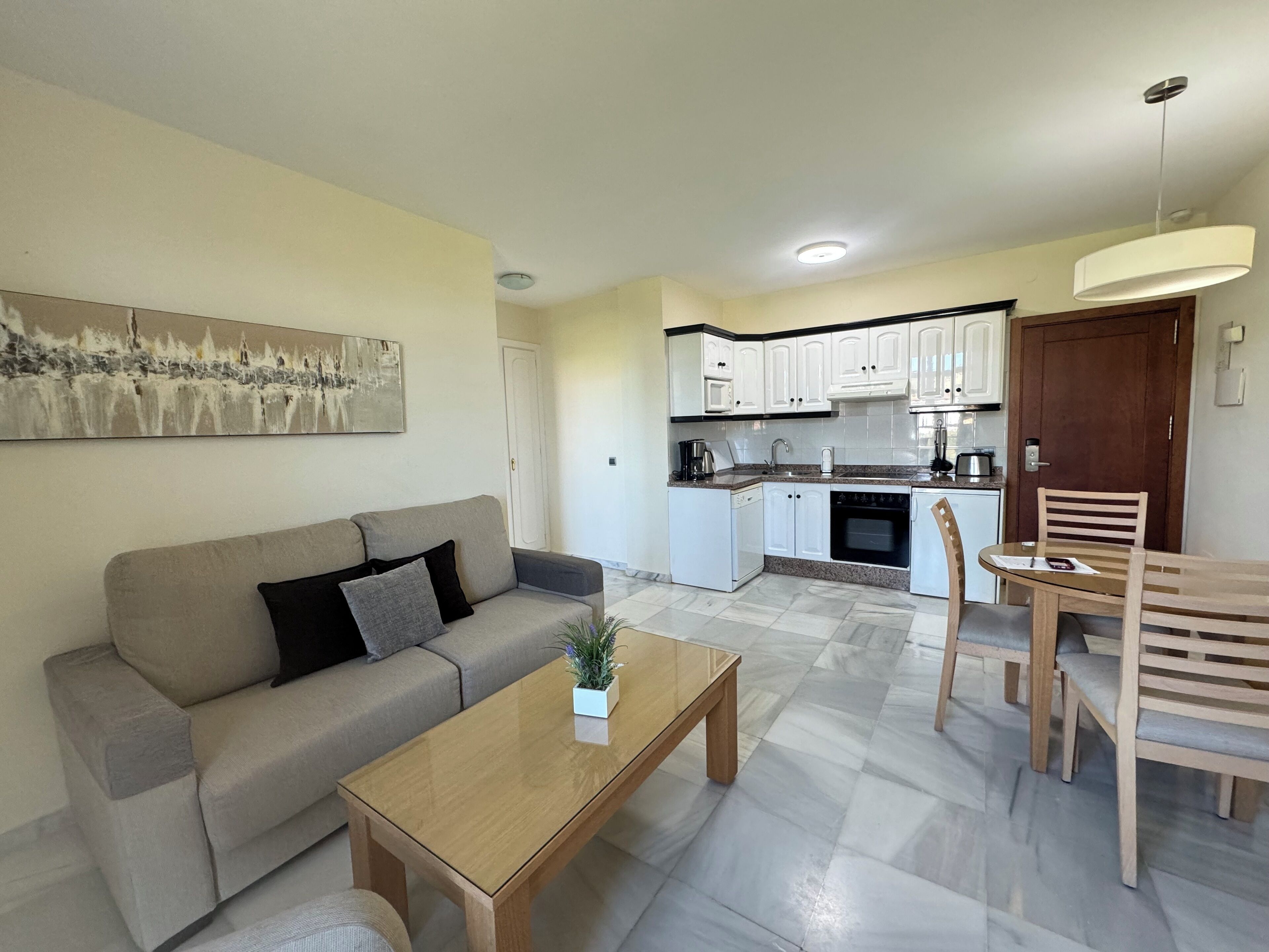 Una sala de estar con un sofá, una mesa de centro y un comedor con mesa y sillas. Se ve una cocina con armarios y electrodomésticos blancos al fondo.