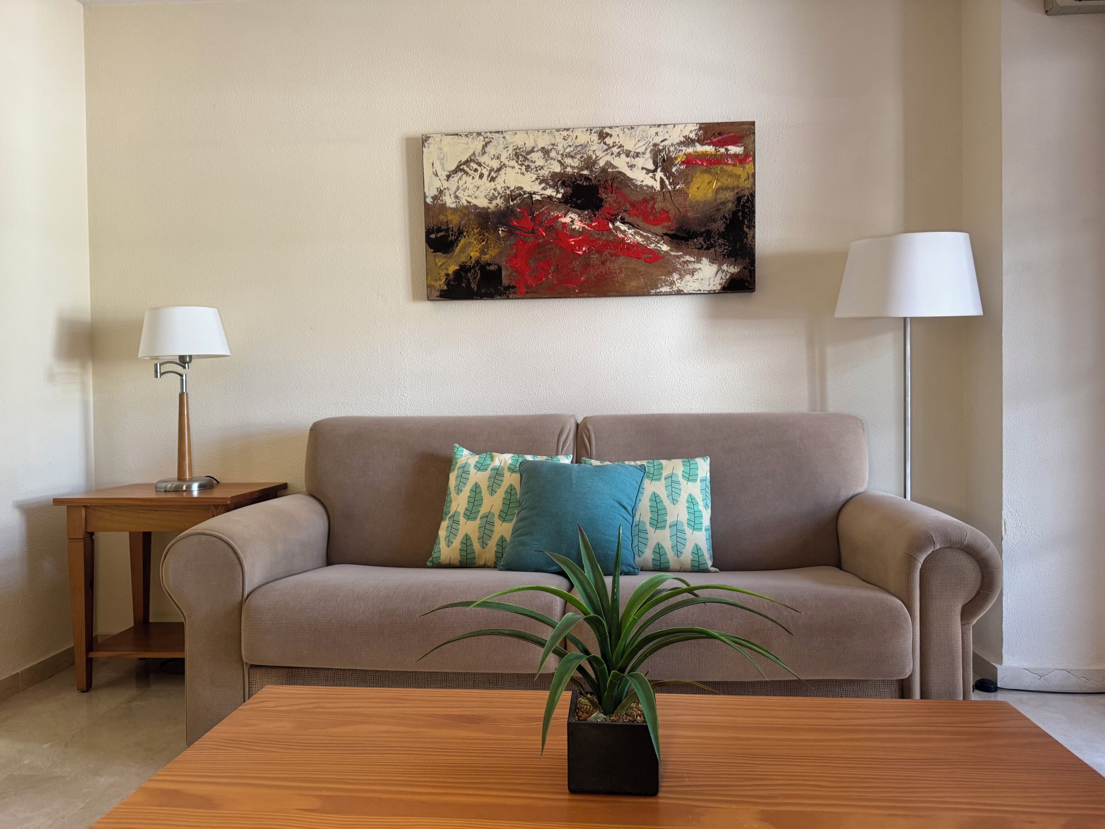 A living room with a beige sofa, a wooden coffee table, a potted plant, a side table with a lamp, and a painting on the wall.