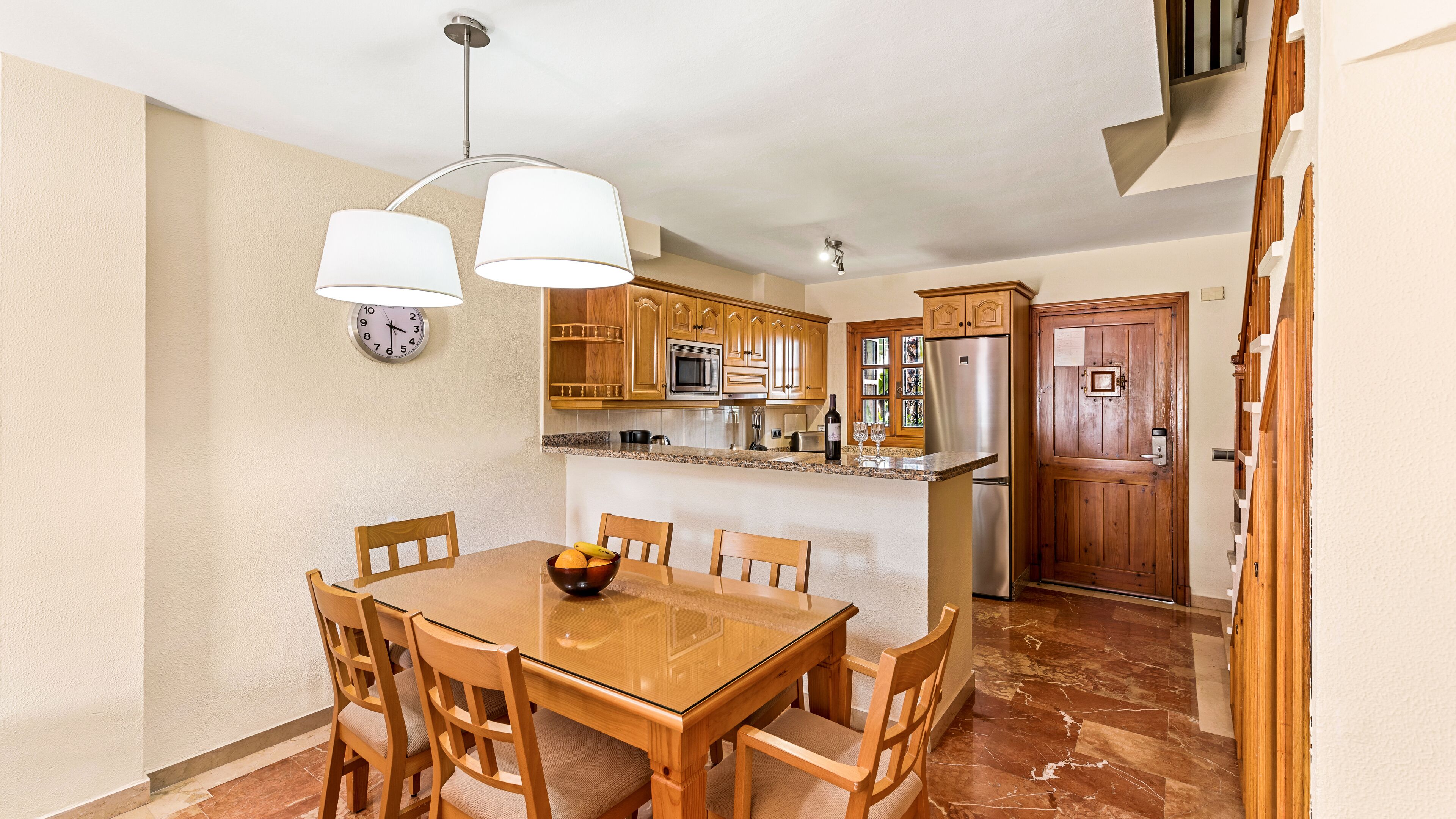 A kitchen with wooden cabinets, a microwave, and a refrigerator. There is a dining table with chairs and a clock on the wall.