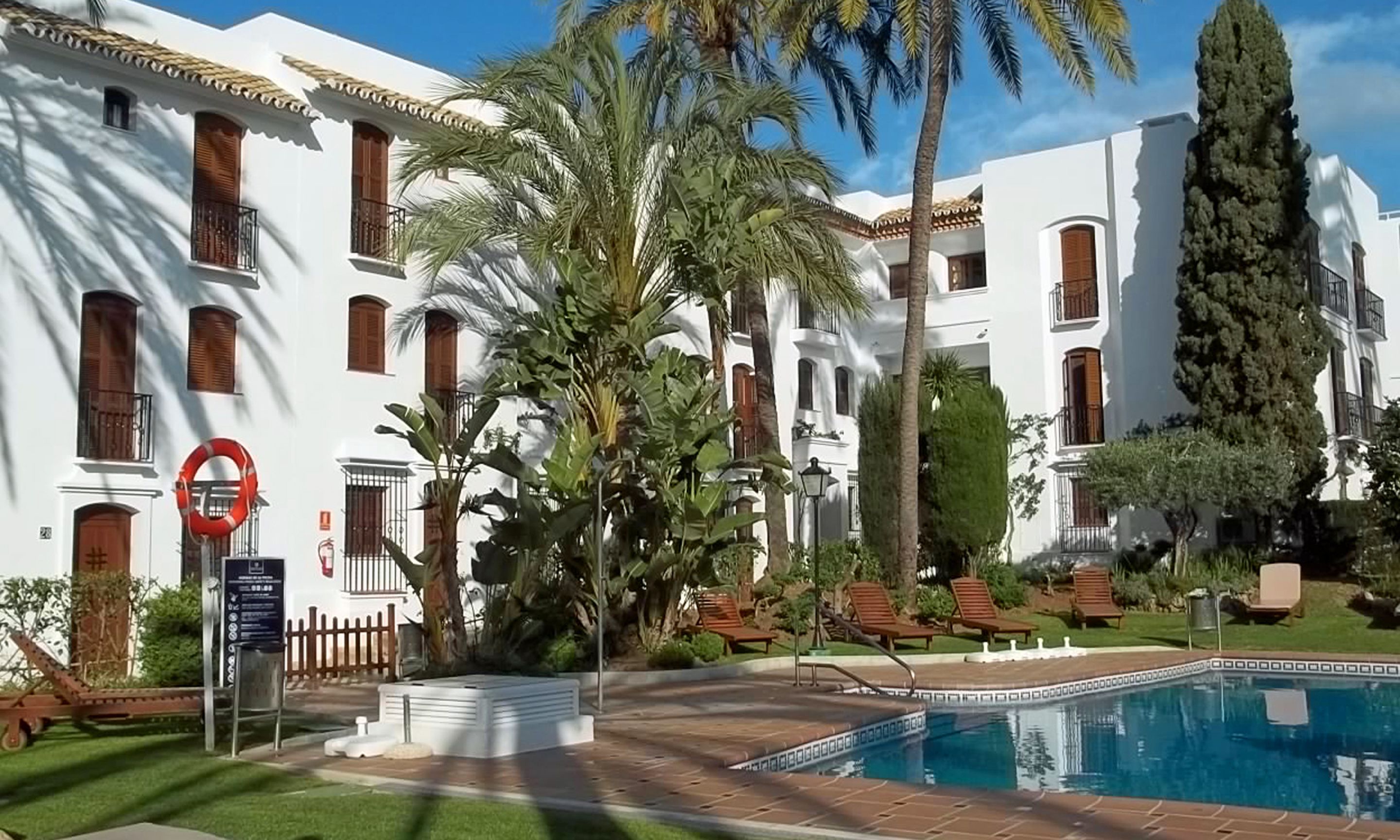 A poolside area with a swimming pool, palm trees, and a white building with balconies.