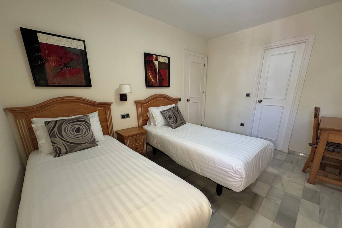 Ett hotellrum med två separata sängar, en träpanel som huvudgavel, ett litet träbord vid sängen, en vägglampa och en dörr.