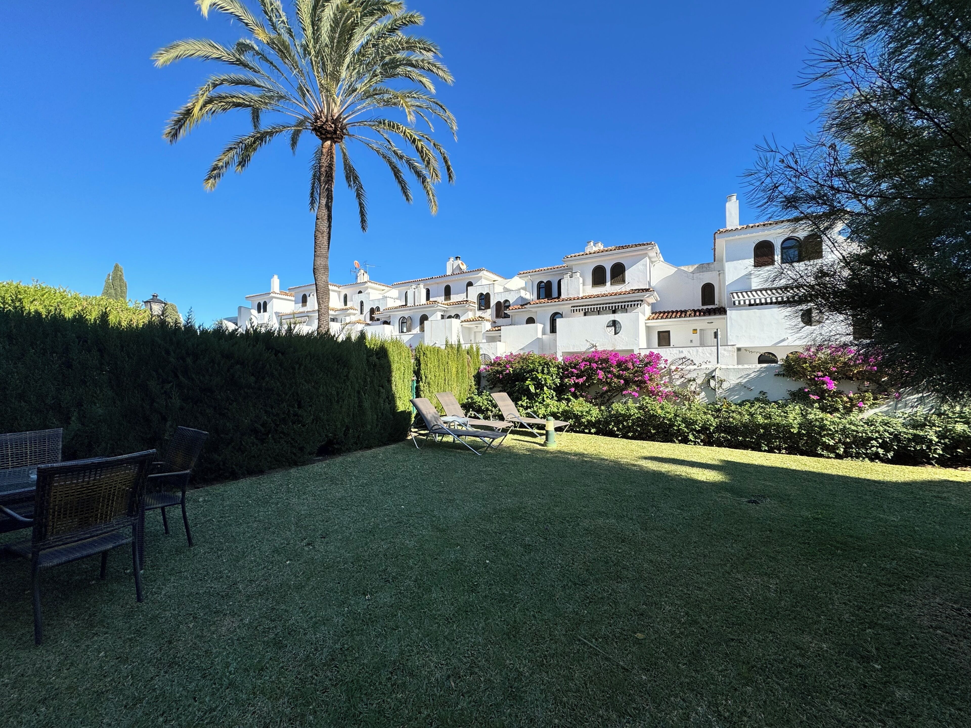 A white villa with multiple balconies, a palm tree, and a garden with pink flowers.