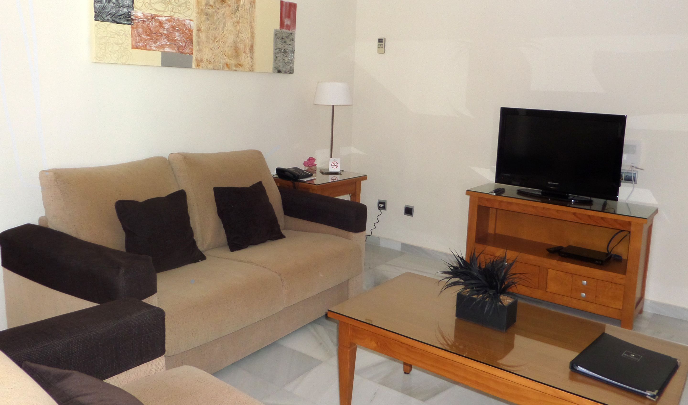 A living room with a beige sofa, a glass coffee table, a wooden TV stand, and a small plant.