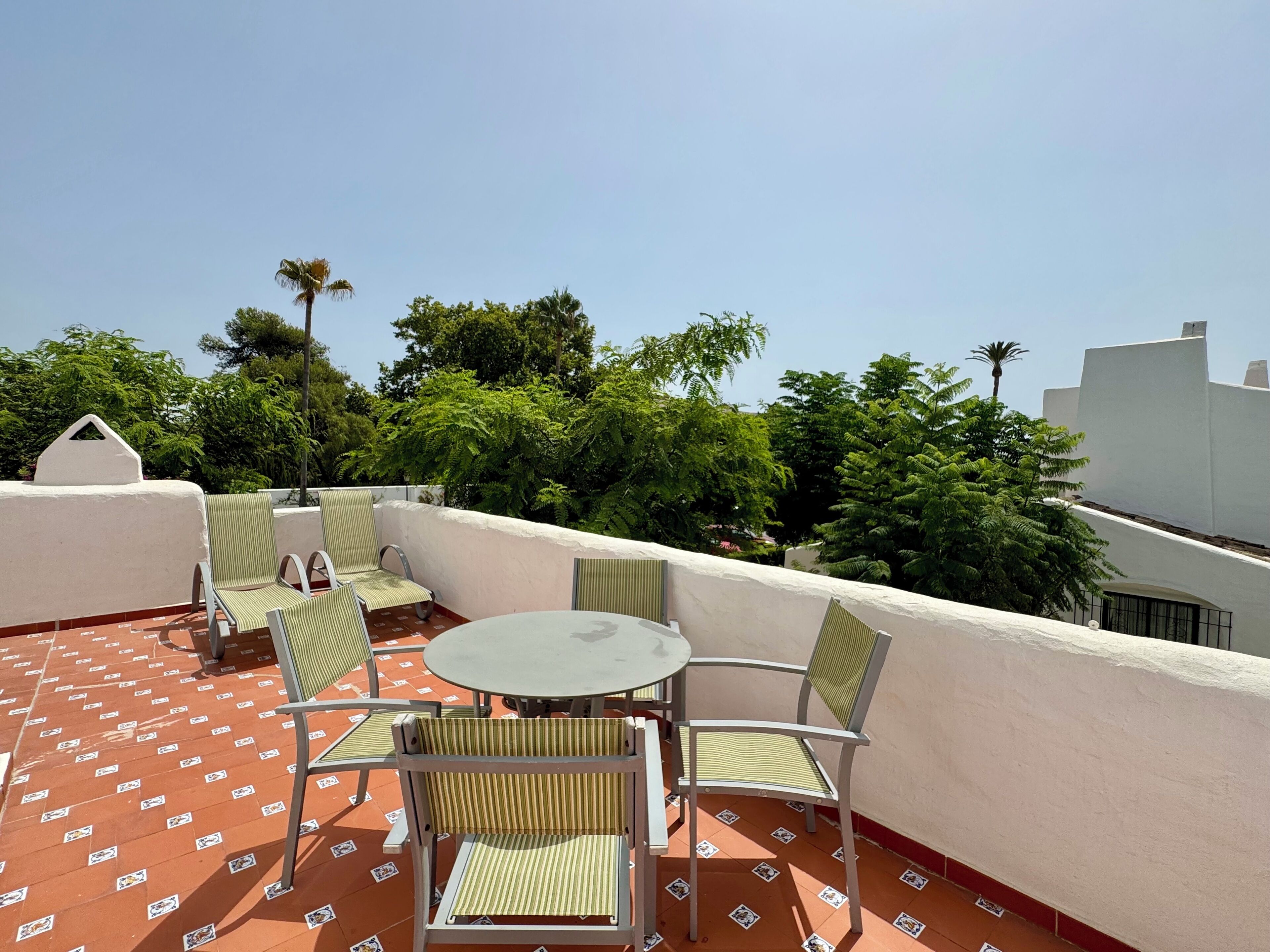 A rooftop terrace with a round table and four chairs, surrounded by a white wall and red-tiled floor.