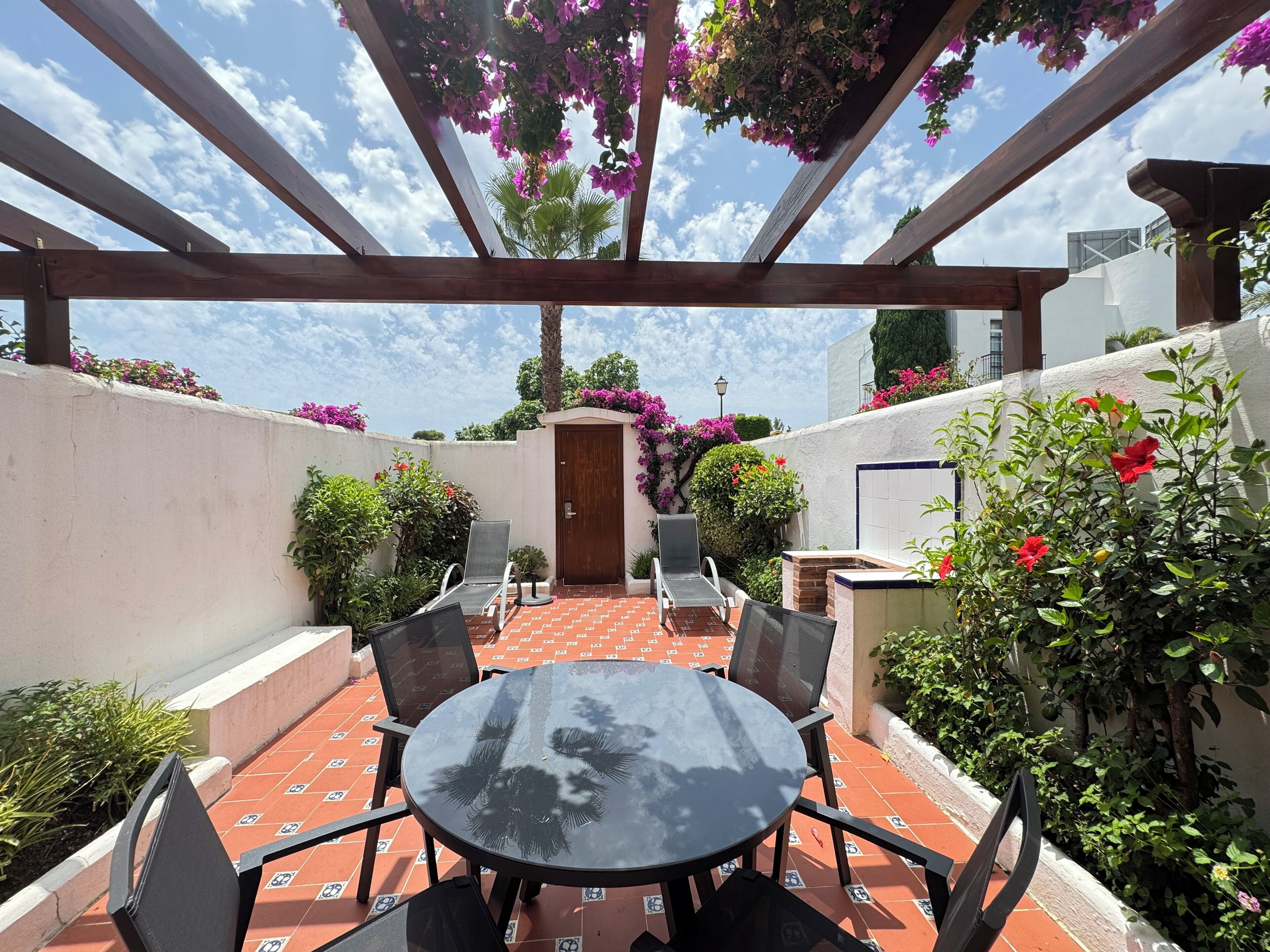 An outdoor patio with a round table and chairs, surrounded by potted plants and flowering shrubs.
