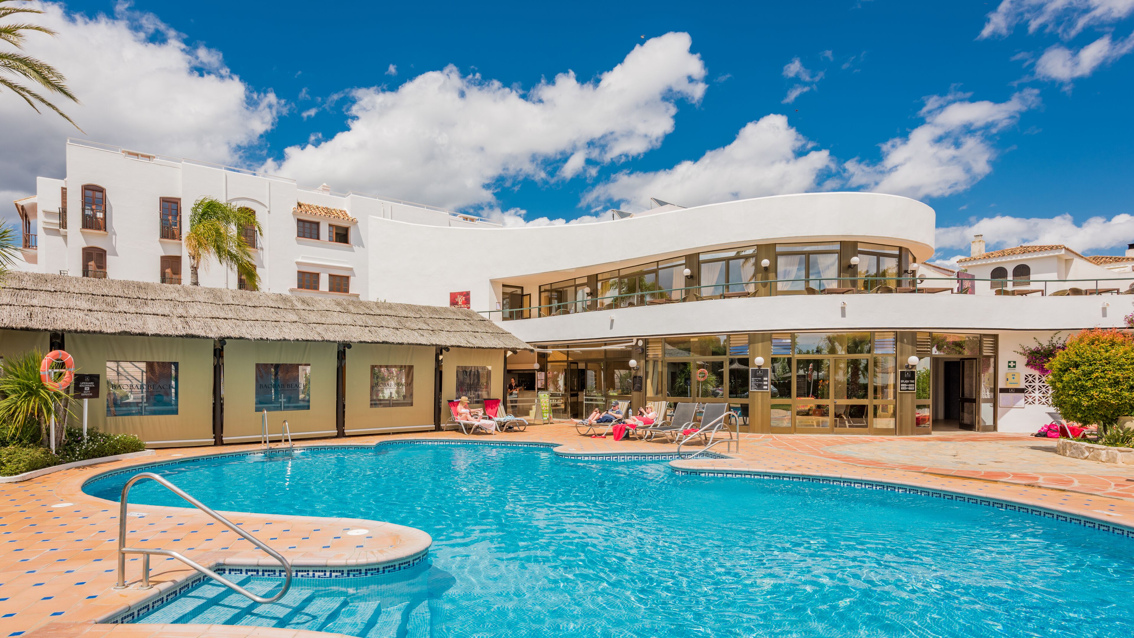 A pool area with a clear blue pool, lounge chairs, and a building with a white facade and a thatched roof structure.