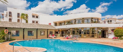 A pool area with a clear blue pool, lounge chairs, and a building with a white facade and a thatched roof structure.
