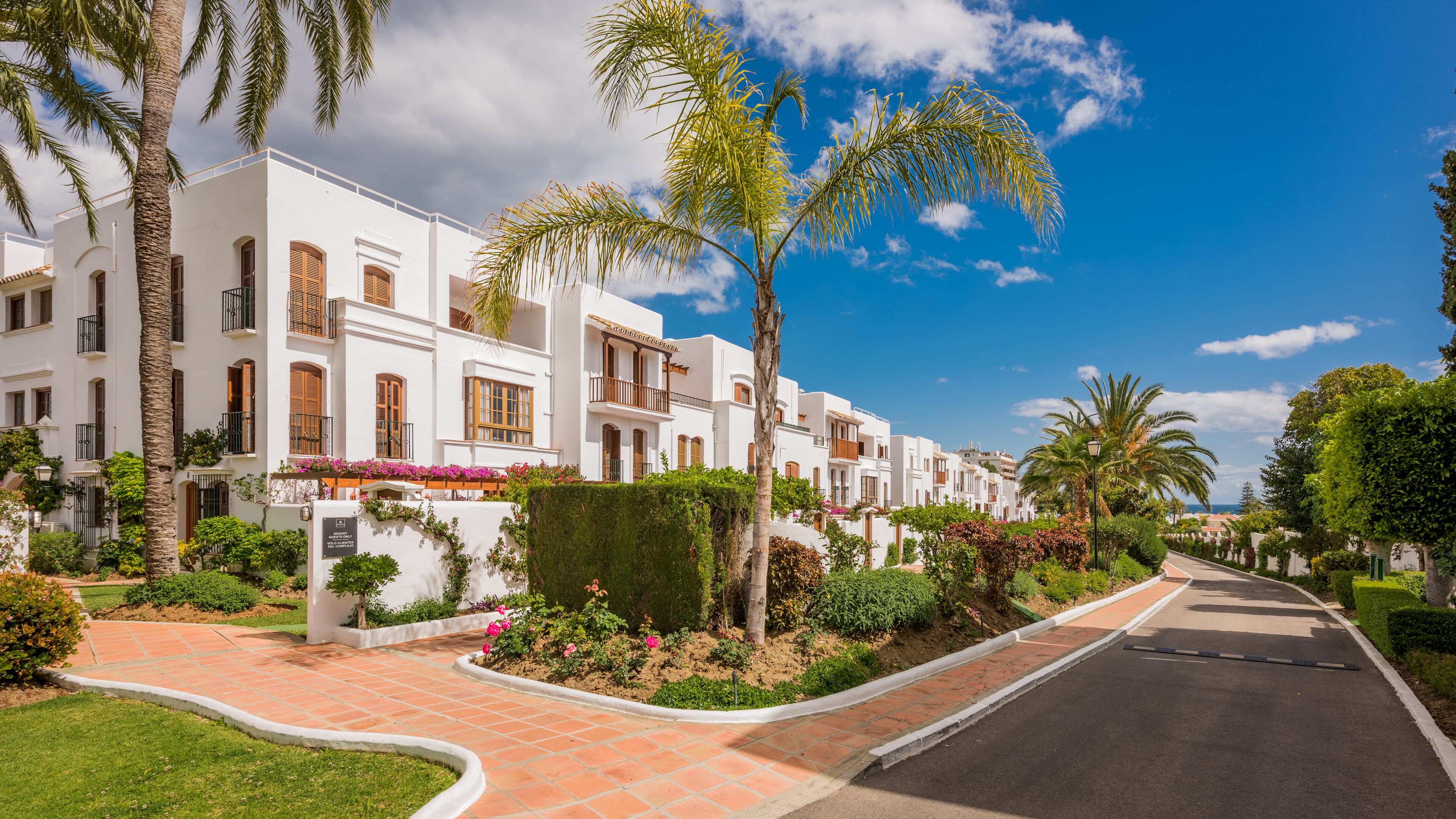 A row of white buildings with balconies, surrounded by palm trees and well-maintained gardens.