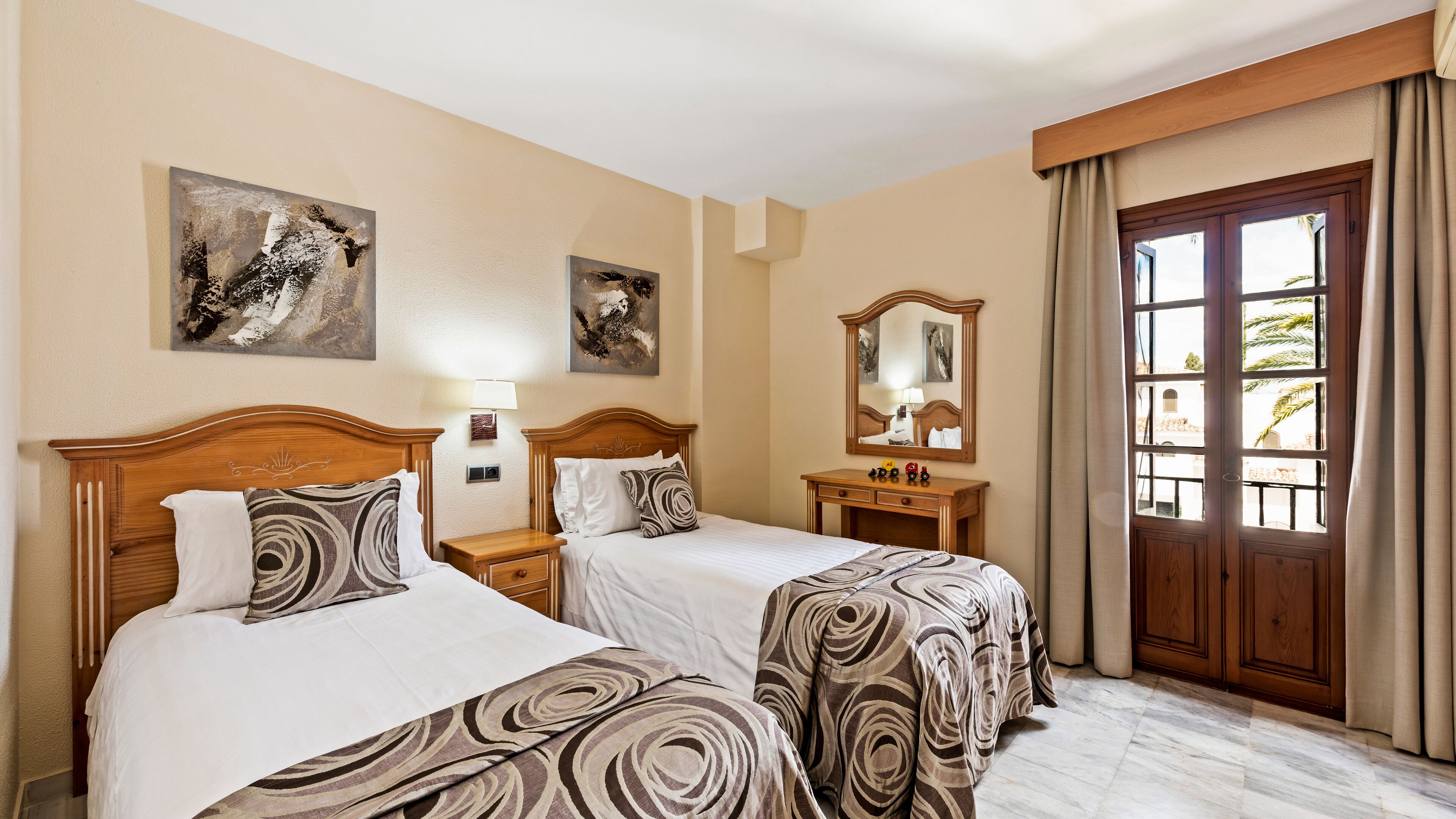 Quarto de hotel com duas camas, cabeceira de madeira, mesa de cabeceira com abajur, cômoda e uma janela grande com cortinas.