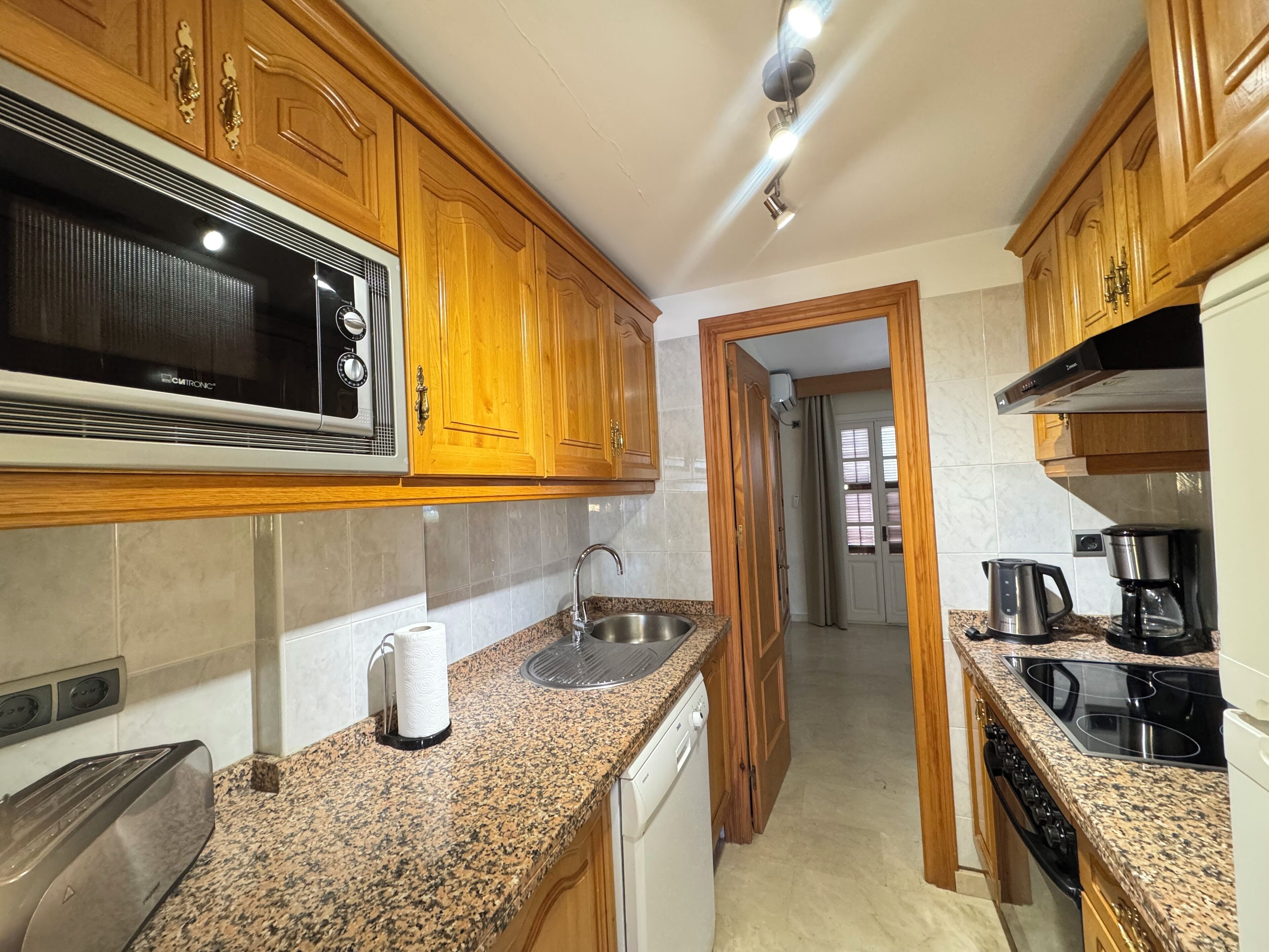 Cocina compacta con armarios de madera, microondas, horno empotrado, cafetera y lavavajillas.