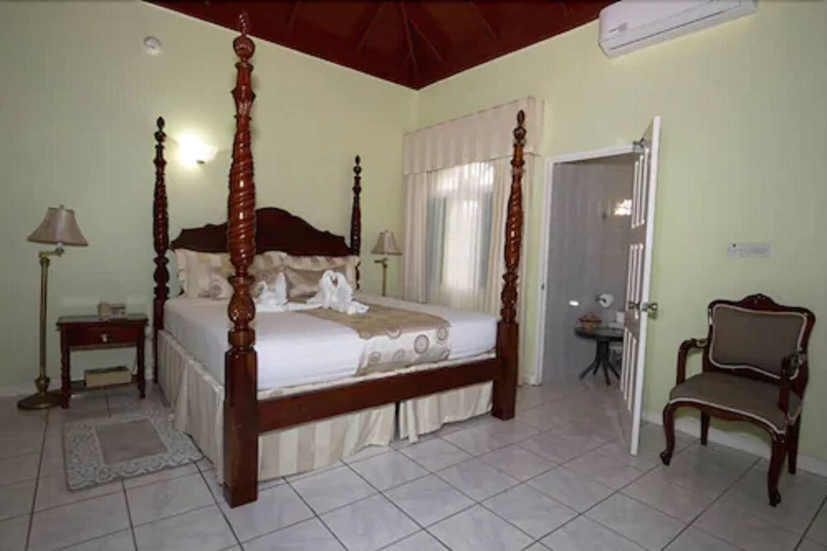 A four-poster bed with a wooden frame, a bedside table with a lamp, a chair, and a door leading to another room.