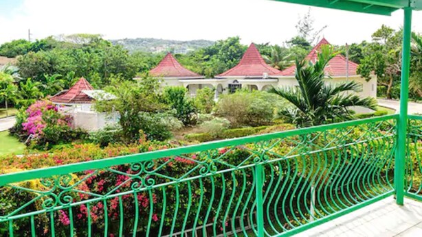 Un balcon doté d’une balustrade verte, donnant sur un jardin où se trouvent des bâtiments aux toits rouges et une végétation luxuriante.