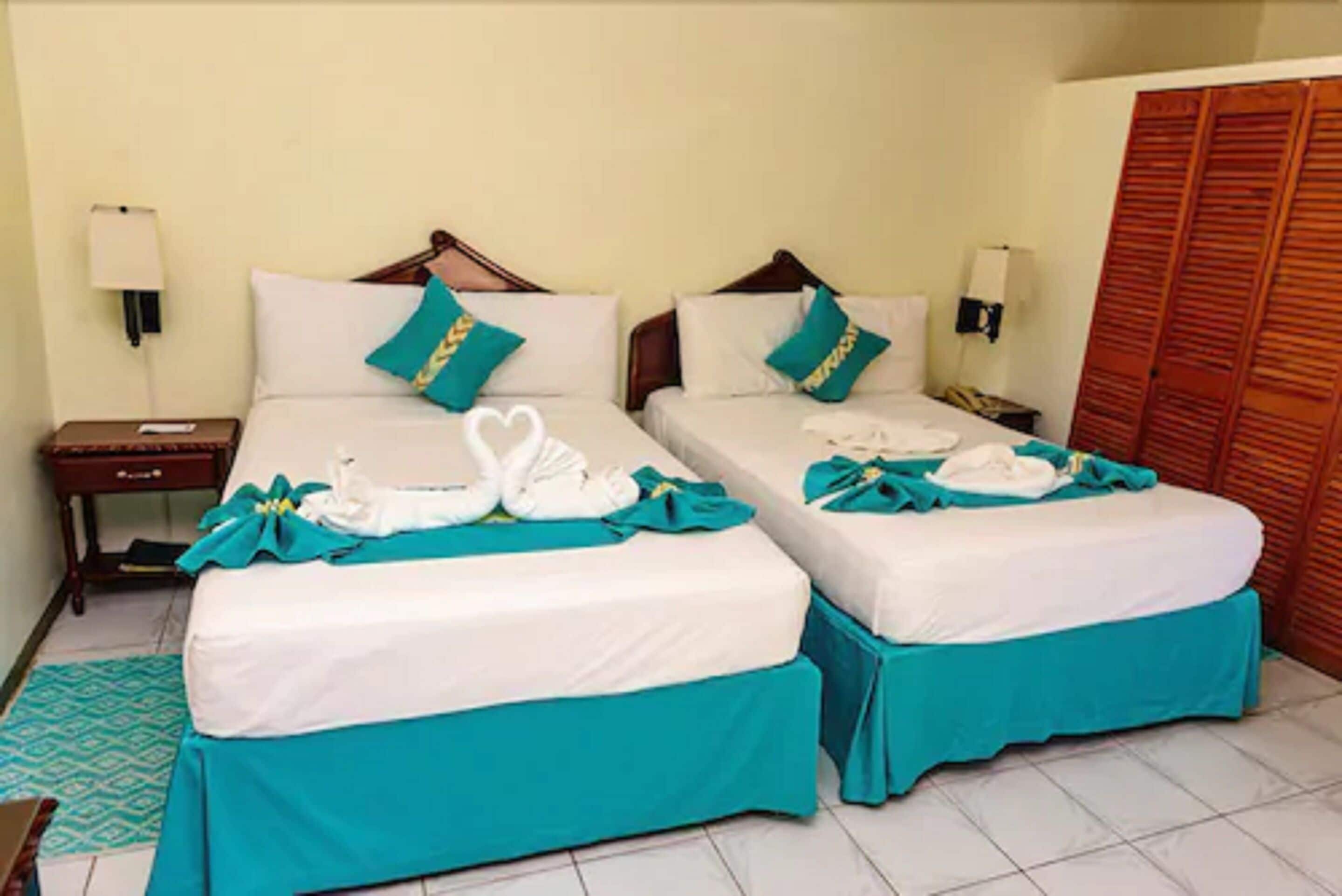 A hotel room with two beds, each with teal bedspreads and white linens. There are two bedside tables with lamps, a wooden wardrobe, and a patterned rug on the floor.