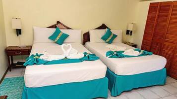 A hotel room with two beds, each with teal bedspreads and white linens. There are two bedside tables with lamps, a wooden wardrobe, and a patterned rug on the floor.