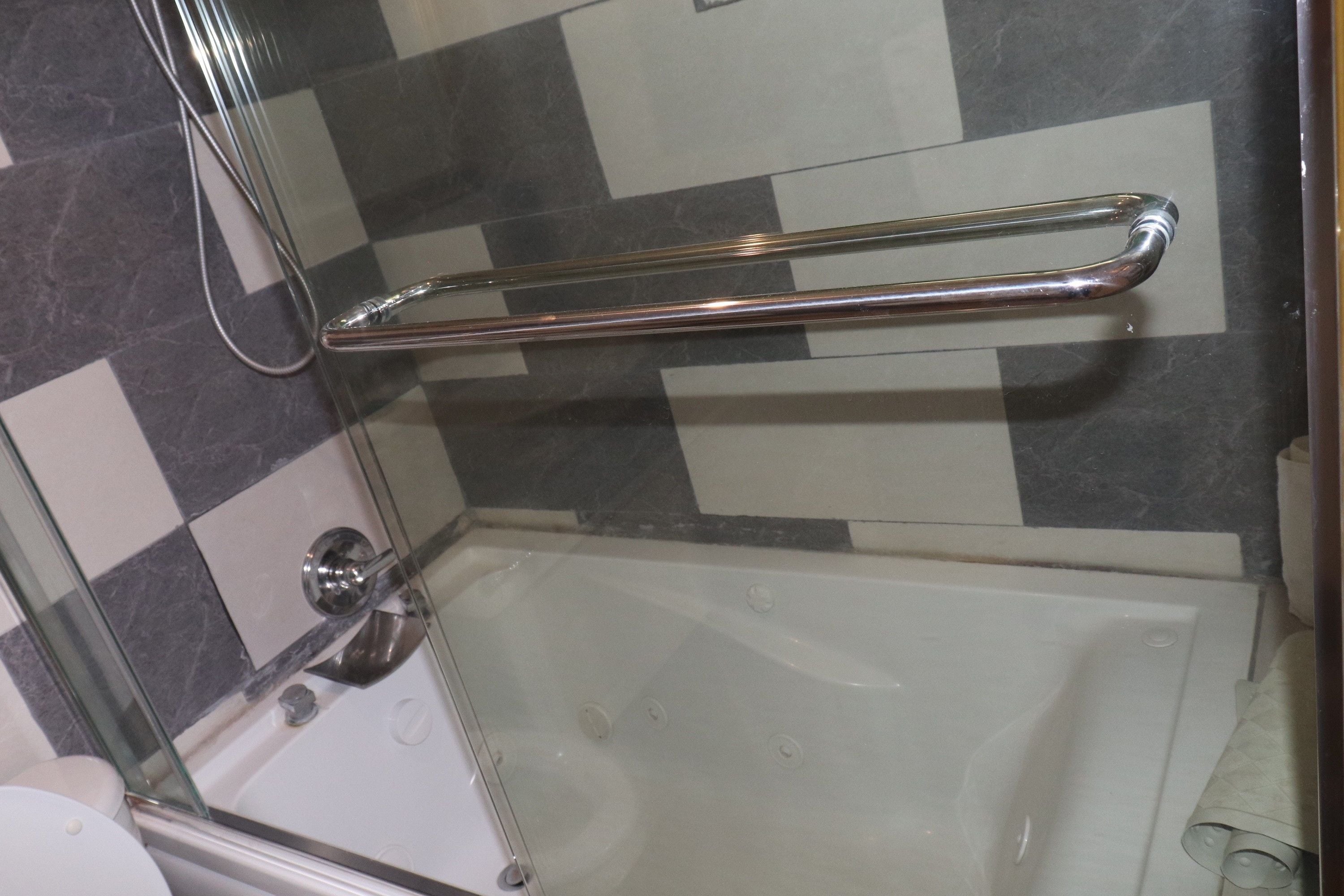 Ducha con cerramiento de vidrio, barra de apoyo cromada, paredes con azulejos de cuadros y una bañera blanca.